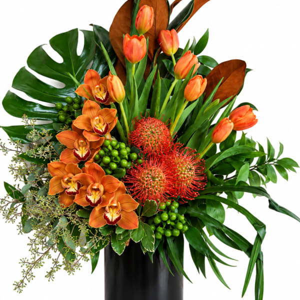 Orange tulips and orchids arranged in a black vase with tropical foliage