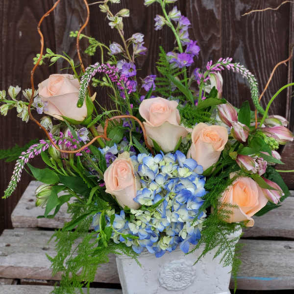 Mixed bouquet with pale roses, blue hydrangea, and purple blooms in a white vase