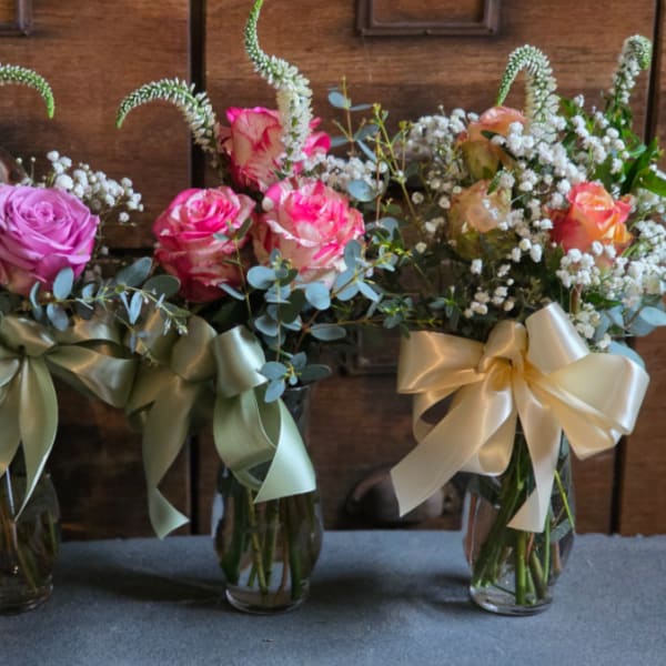 Three small glass vases with pink and peach roses, white fillers, and large satin ribbon bows.