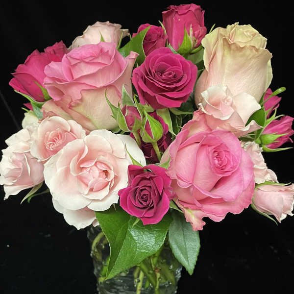 Pink and blush roses arranged tightly in a small clear glass vase against a black background