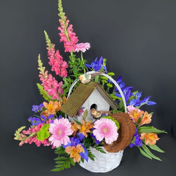 Basket arrangement with pink snapdragons, gerbera daisies, blue irises and a birdhouse centerpiece