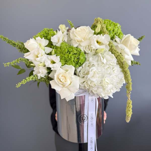 White and green flower arrangement with roses and hydrangeas in a tall silver hatbox-style container