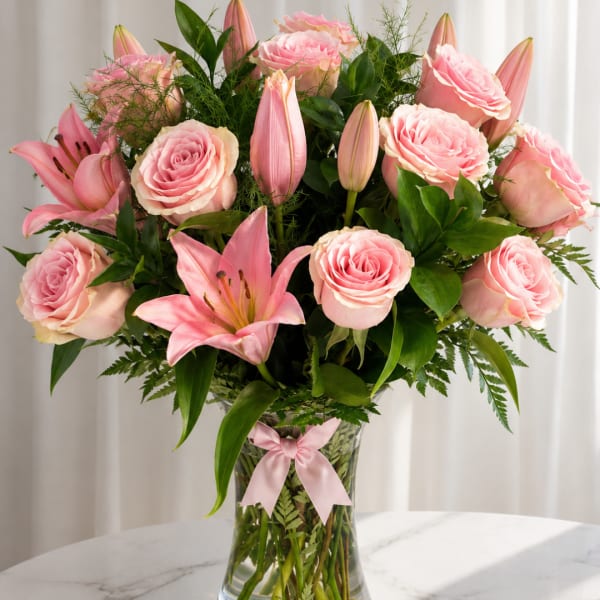 Pink roses and lilies arranged in a clear glass vase with a pink ribbon