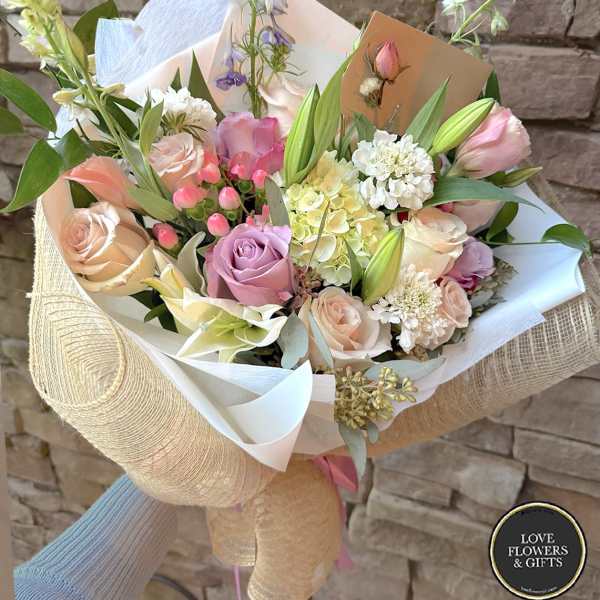 Hand-tied bouquet of pastel roses, lilies, and mixed blooms wrapped in white and burlap-style paper