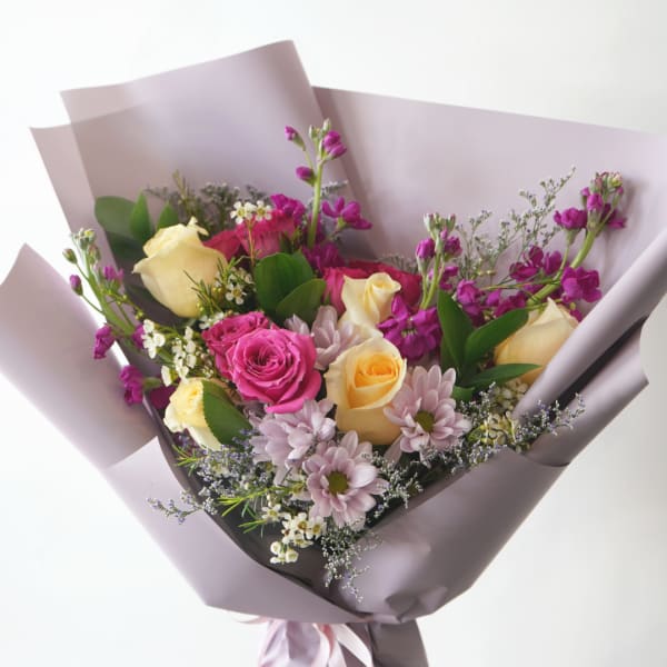 Hand-tied bouquet of yellow and pink roses with purple flowers wrapped in lavender paper.