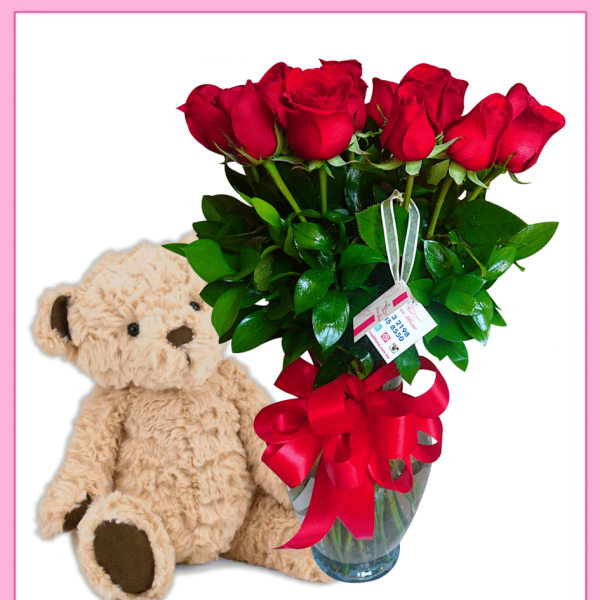 Red roses in a glass vase beside a teddy bear