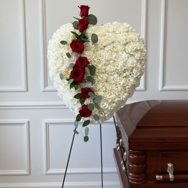Heart-shaped white floral spray with red roses on a stand beside a casket