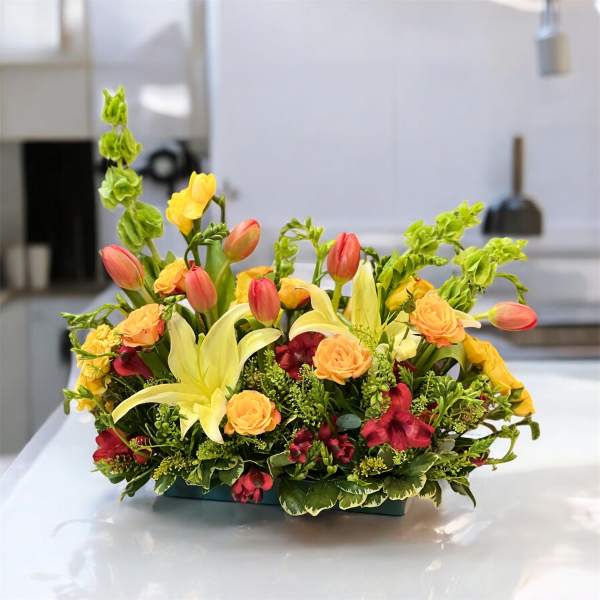 Mixed floral arrangement with yellow lilies, orange roses, and red tulips in a low container