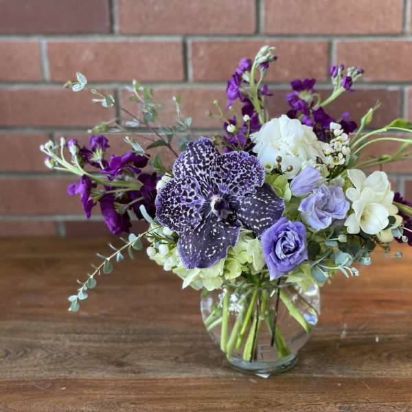 Low glass vase with purple orchid, lavender lisianthus, white roses and mixed blooms on a table.