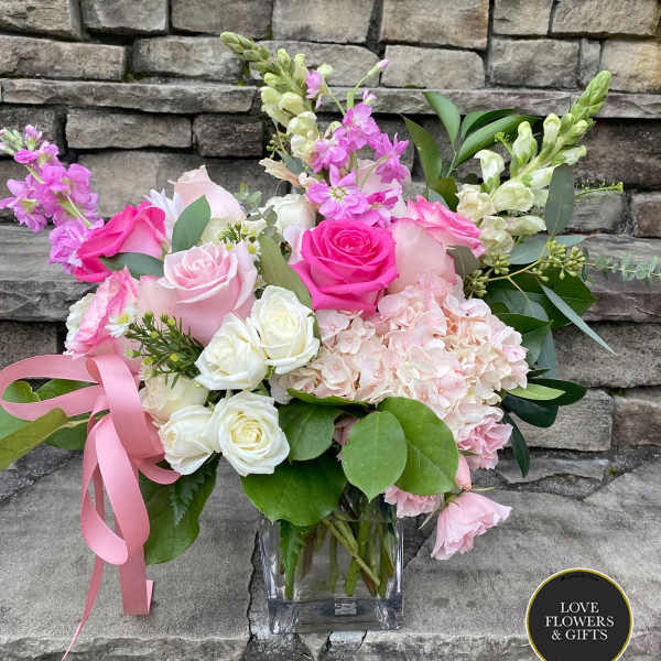 Pink and white roses, hydrangeas, and snapdragons in a clear glass cube vase with a pink ribbon.