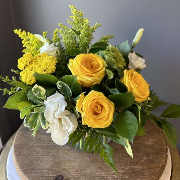 Compact arrangement of yellow roses with white blooms and fillers in a clear glass vase