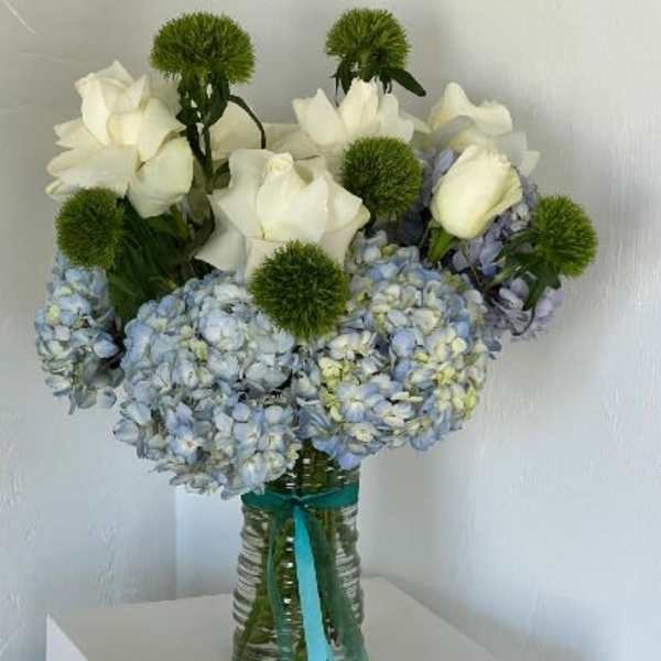White roses and blue hydrangeas in a glass vase with green pom-pom accents