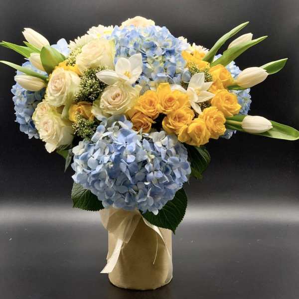 Blue hydrangeas with yellow and white roses in a wrapped vase arrangement