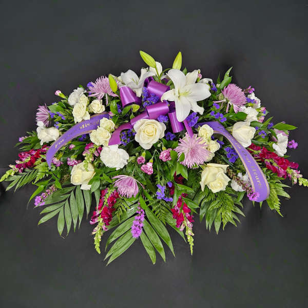 Elongated spray of white lilies, roses and purple flowers with a purple sympathy ribbon on a dark background