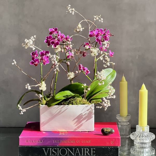 Pink orchid arrangement in a white rectangular planter beside two yellow candles