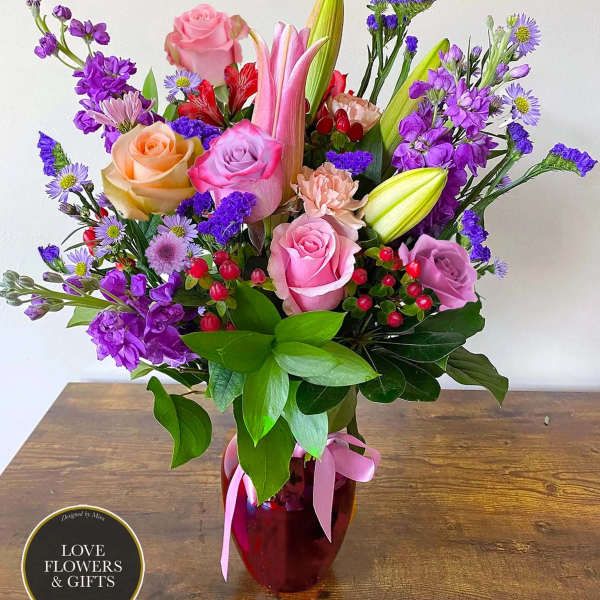 Mixed bouquet of pink roses, lilies, and purple flowers in a red glass vase with pink ribbon.