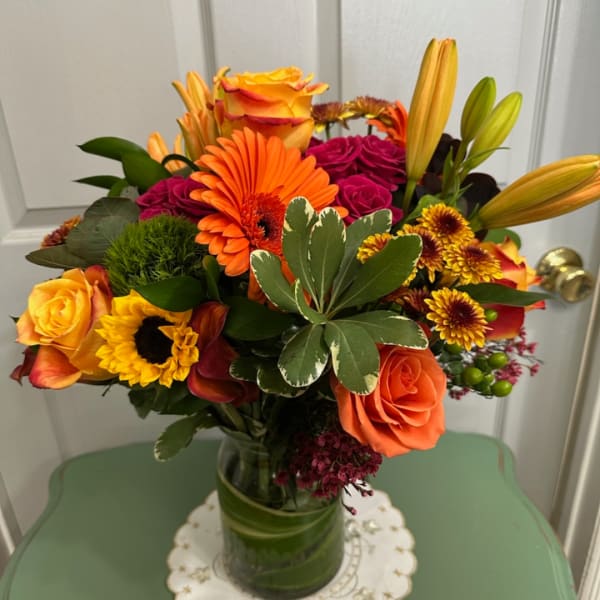 Bright mixed bouquet of roses, lilies, gerbera daisies, and sunflowers in a glass vase