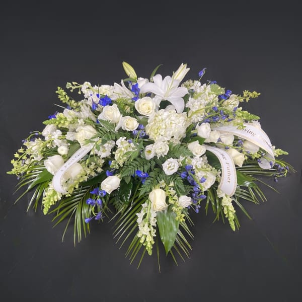 Large white and blue floral spray with roses, lilies, and palm fronds accented by memorial ribbons.