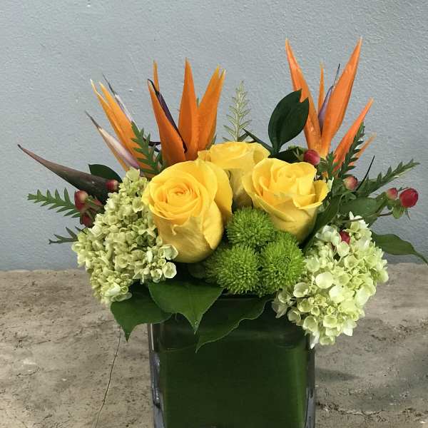 Yellow roses and green hydrangeas in a square glass vase with orange tropical accents