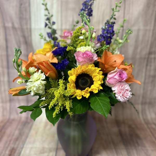 Mixed bouquet in a tall vase with sunflowers, roses, lilies, and purple spikes