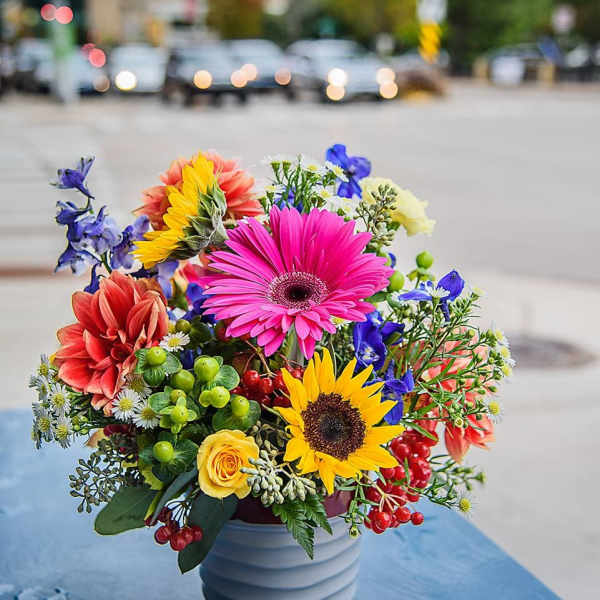 Bright mixed bouquet with sunflower, pink gerbera, and roses in a gray pot on a blue table outdoors.