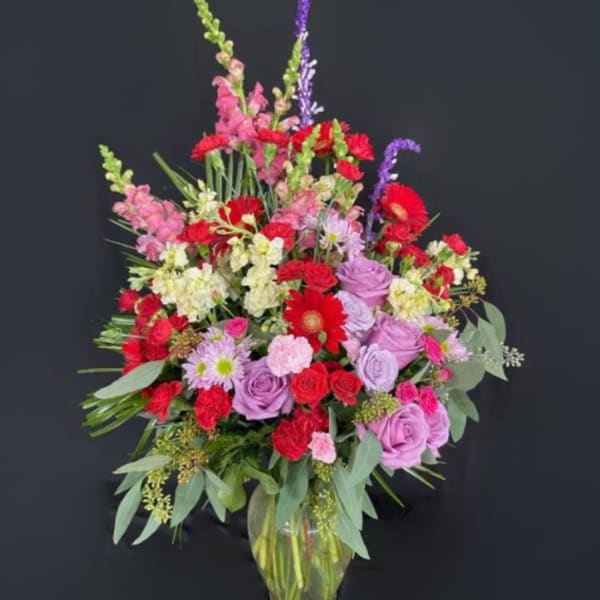 Tall mixed arrangement of red, pink, yellow, and lavender flowers in a clear glass vase