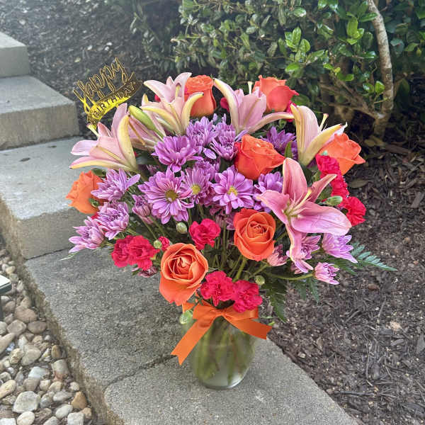Mixed bouquet of pink lilies, orange roses, and purple daisies in a glass vase