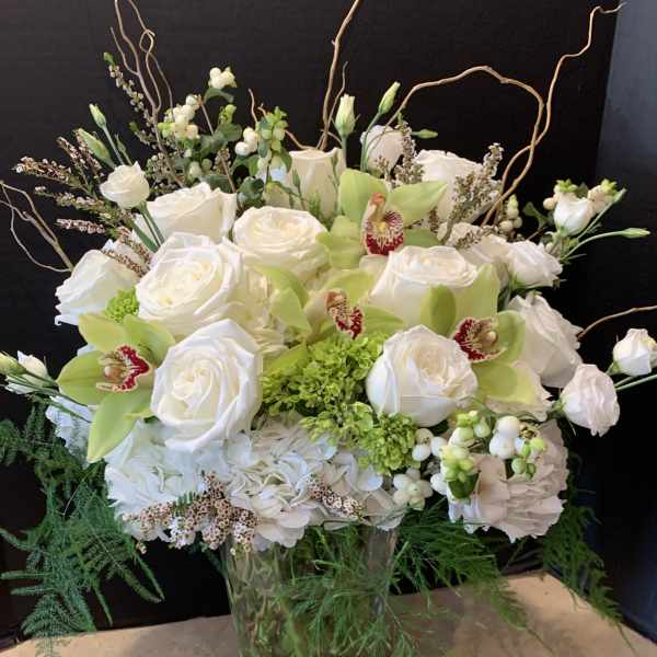 Tall white and green arrangement with roses, hydrangea, and cymbidium orchids in a clear glass vase