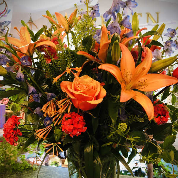 Tall mixed arrangement with orange lilies, an orange rose, red blooms and blue delphinium in a clear glass vase