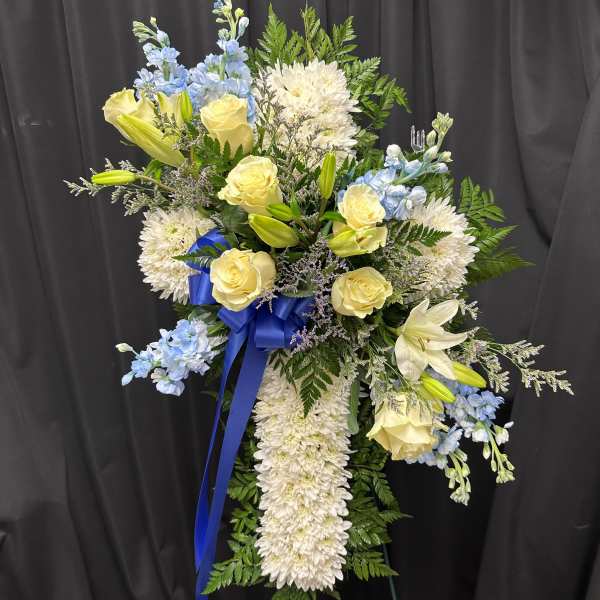Standing spray of white mums, yellow roses, lilies, and blue flowers with blue ribbon on an easel.