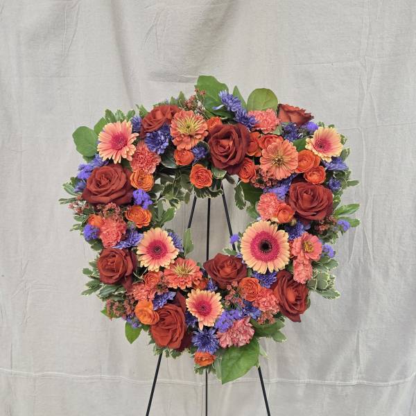Heart-shaped standing wreath with red roses, orange gerbera daisies, and purple flowers on a black easel