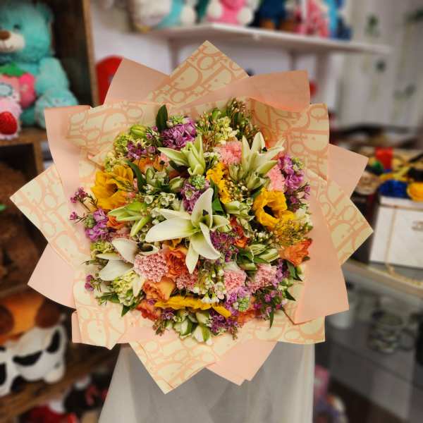 Mixed bouquet of lilies, sunflowers, carnations, and purple blooms in peach wrap