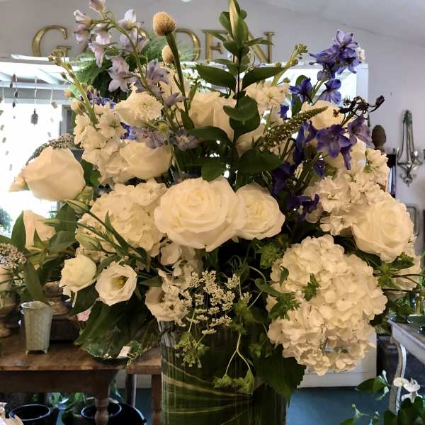Tall arrangement of white roses and hydrangeas with purple delphinium in a clear glass pedestal vase