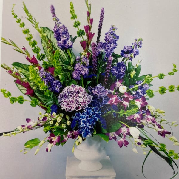 Tall arrangement of purple and blue hydrangeas, stock, orchids, and bells of Ireland in a white pedestal vase
