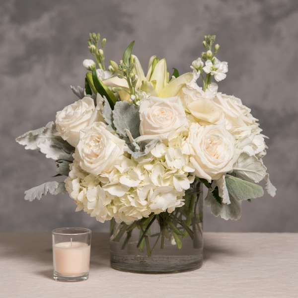 White roses and hydrangeas in a glass vase with a small candle beside it