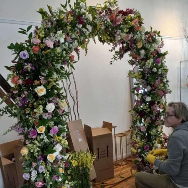 Large floral arch covered in mixed blooms with a person arranging it nearby