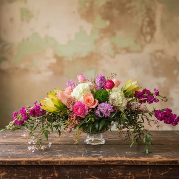 Low centerpiece of pink and peach roses with white hydrangeas, ranunculus and magenta stock in a clear glass bowl.