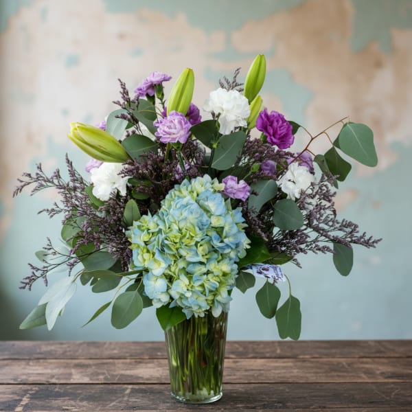 Mixed bouquet with blue hydrangea, purple and white blooms, and lily buds in a clear glass vase.