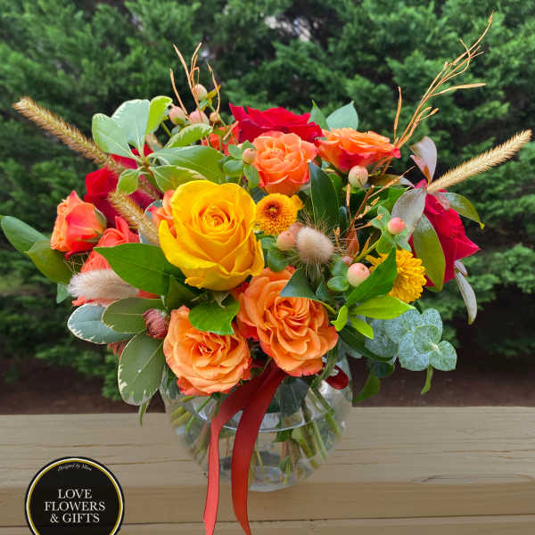 Round glass vase of yellow, orange, and red roses with small yellow mums and berries, tied with an orange ribbon