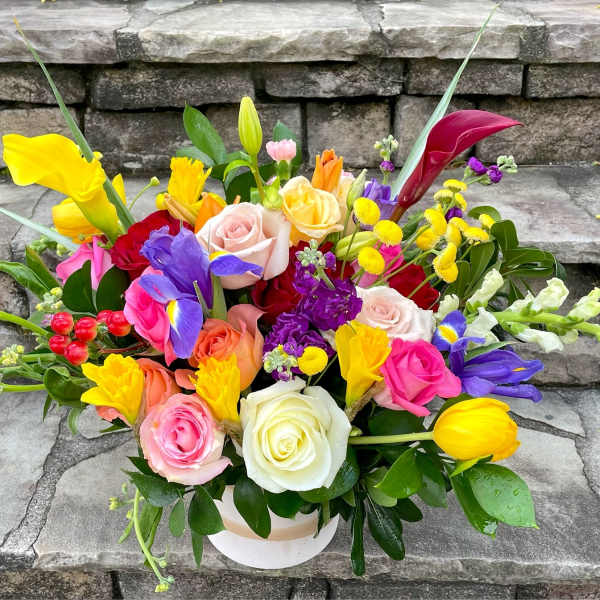 Low round arrangement of colorful roses, tulips, irises, and other blooms in a white container.