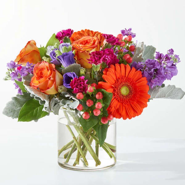 Bright mixed bouquet of orange gerbera, roses, and purple blooms in a clear glass cylinder vase