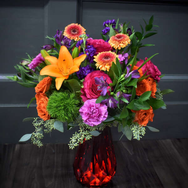 Bright mixed bouquet in a red glass vase with orange, pink, and purple flowers.