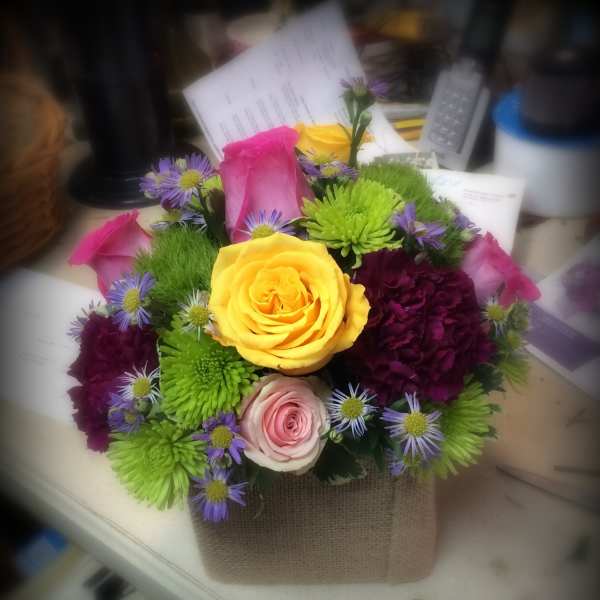 Mixed bouquet with yellow, pink, and purple flowers in a burlap-wrapped container