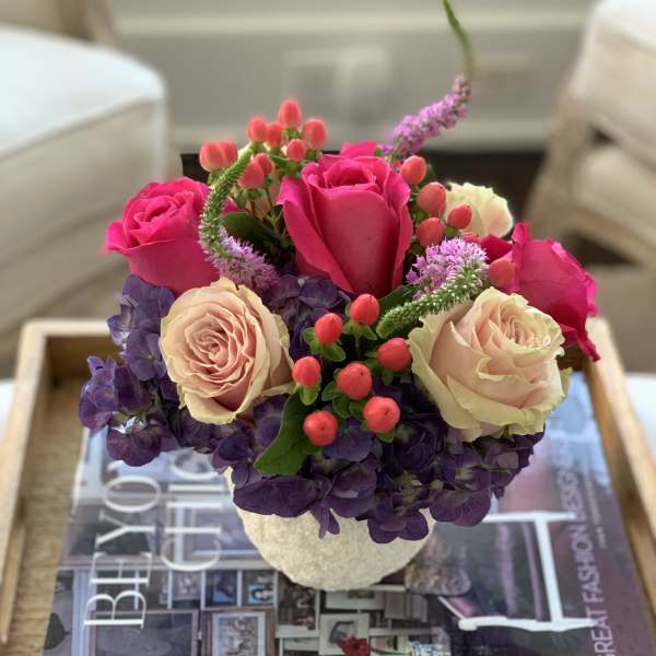 Pink and cream roses arranged in a small white vase with purple flowers.