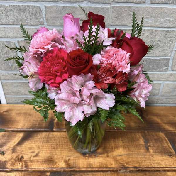 Pink and red roses, carnations, and alstroemeria arranged in a clear glass vase on a wooden table.