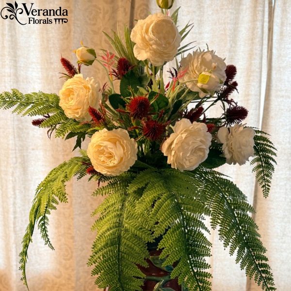 Tall arrangement of cream blooms with red accents and cascading fern fronds in a red ceramic vase