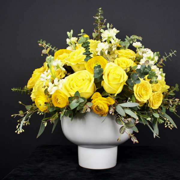 Yellow roses and ranunculus in a white pedestal vase