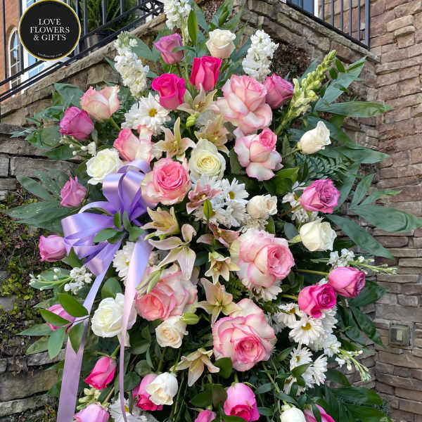 Tall standing spray of pink and white roses, lilies, and daisies with a lavender ribbon on an easel