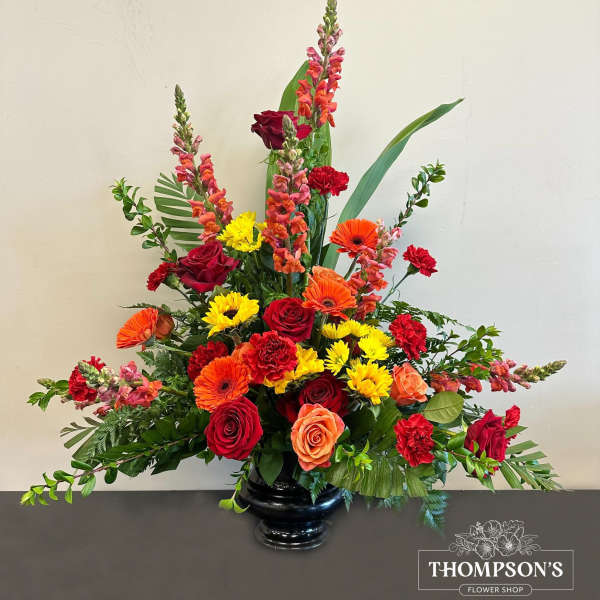 Tall mixed arrangement of red roses, orange gerbera daisies, yellow mums and snapdragons in a black vase.