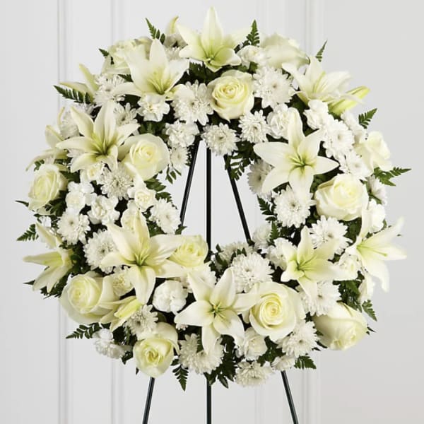 White floral wreath on a black stand with lilies and roses